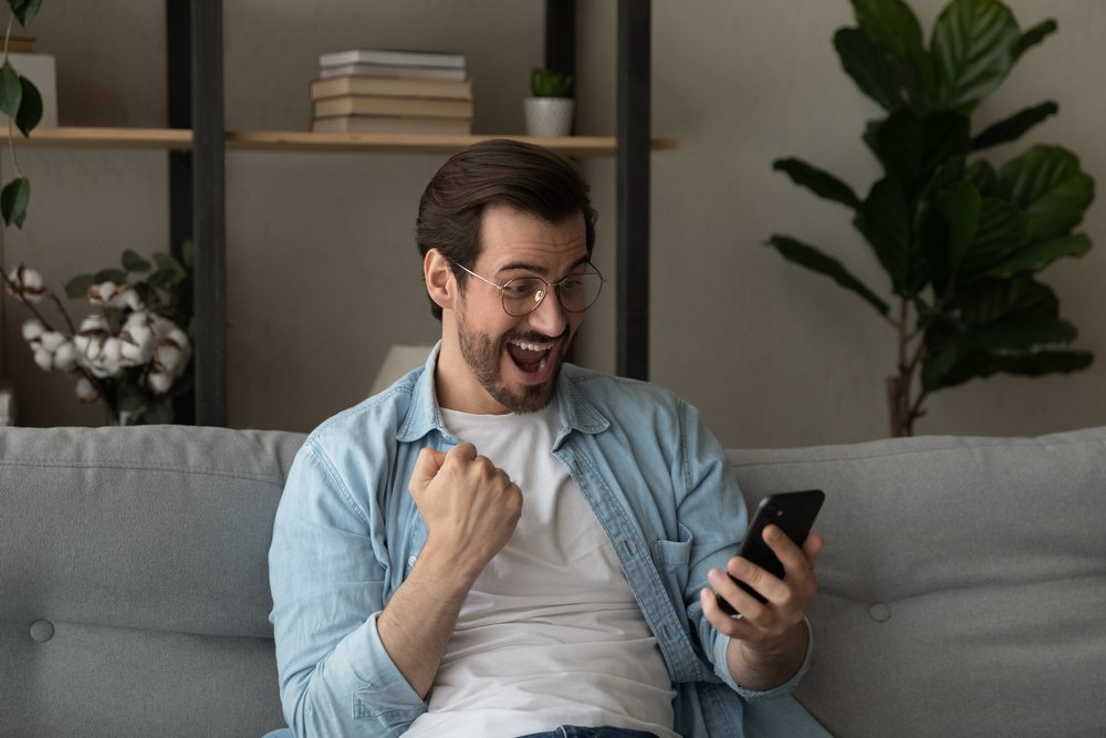 Excited,overjoyed,young,man,sit,on,sofa,holding,smartphone,feeling